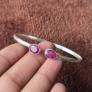 Könnte beinhalten: Ein offenes Armreif-Armband in Silberoptik mit zwei oval geschliffenen Rubin-Edelsteinen. Die Rubine sind in einer Zargenfassung gefasst, und das Armband hat eine glatte, polierte Oberfläche. Die Edelsteine sind tiefrot.