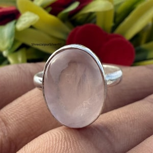 Könnte beinhalten: Ein Silberring mit einem großen, ovalen Rosenquarz-Edelstein. Der hellrosa Edelstein ist in einem schlichten Silberband gefasst. Der Ring wird vor einem verschwommenen Hintergrund aus Grün und Rot gezeigt.