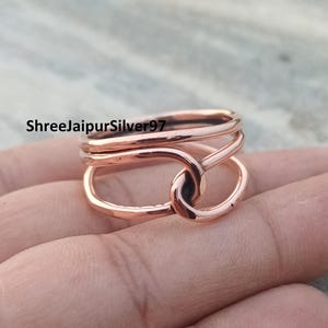 Op de afbeelding: Een roségouden ring met een knoopontwerp. De ring heeft een reeks gebogen banden die in elkaar grijpen om een knoop in het midden te vormen. De ring is glad en glanzend. De tekst "ShreeJaipurSilver97" is zichtbaar.