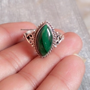 May include: A silver ring with a striking, marquise-shaped malachite gemstone. The vivid green stone is framed by a rope-style bezel. The ring has decorative scrollwork on the shoulders. The text "ShreejaipurSilver97" is visible on the ring.