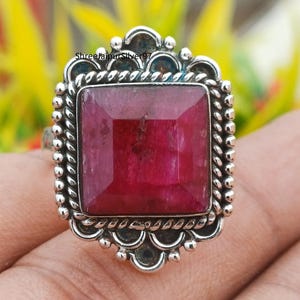 May include: A silver ring featuring a large, square-cut ruby gemstone. The ruby is a deep red color and is set in a decorative, ornate silver setting. The ring has a textured, rope-like border and a detailed design around the gemstone.