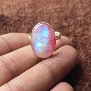 Puede incluir: Anillo de plata con una gran piedra lunar ovalada. La gema presenta una mezcla de colores rosa, azul y blanco, con un efecto brillante. El anillo se muestra en una mano, destacando el tamaño y las variaciones de color de la piedra.