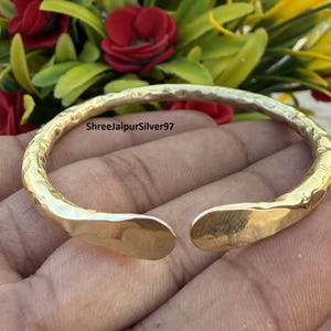 Puede incluir: Una pulsera de puño abierta de color dorado con una textura martillada. La pulsera tiene forma redondeada con extremos ligeramente aplanados y curvados. El fondo incluye flores rojas y amarillas.