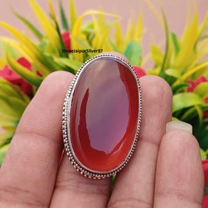 May include: Oval-shaped, reddish-orange gemstone ring with a silver-coloured, textured band. The ring is held in a hand, with the text "ShreejaipurSilver97" visible in the background. The gemstone has a polished, glossy appearance.