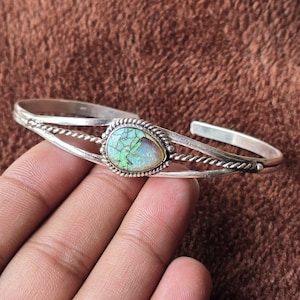 May include: A silver cuff bracelet featuring a pear-shaped, iridescent gemstone. The gemstone is set in a beaded bezel and surrounded by a twisted rope design. The bracelet has a simple, open design with a polished finish.