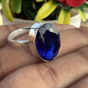Könnte beinhalten: Ein Silberring mit einem großen, facettierten, ovalförmigen, tiefblauen Edelstein. Der Ring hat ein schlichtes Band und eine Zargenfassung. Der Text "ShreeJaipurSilver97" ist auf dem Ring zu sehen.