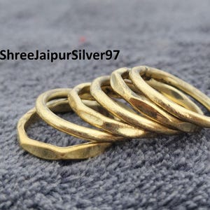 Peut inclure: Une pile de six bagues dorées à la texture martelée. Les bagues sont disposées en ligne légèrement incurvée, avec le texte "ShreeJaipurSilver97" en haut à gauche. Sur une surface grise texturée.