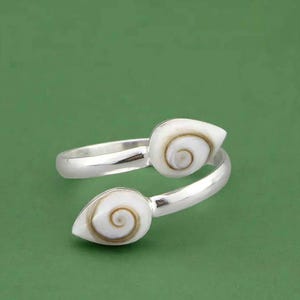 May include: A silver bypass ring featuring two white Shiva eye shell accents. Each shell has a natural brown spiral pattern centred on a smooth, teardrop-shaped white surface. The polished silver band is open-ended, allowing for adjustable sizing.
