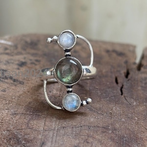 May include: A silver ring featuring three round gemstones. The central stone is a dark, translucent color, while the top and bottom stones are a lighter, opalescent hue. The ring has a unique, open design with curved silver accents.