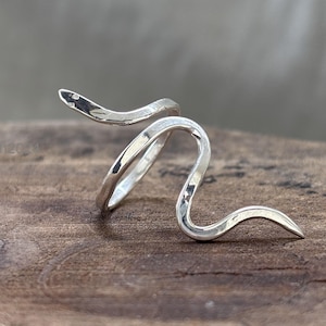 Puede incluir: Anillo plateado con forma de serpiente. El anillo se enrolla alrededor del dedo con un diseño curvo y texturizado. La cabeza de la serpiente está ligeramente levantada y la cola termina en punta. El anillo está sobre una superficie de madera.