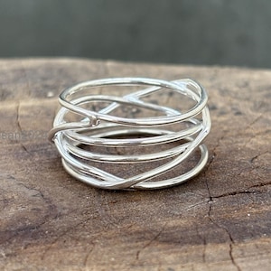 May include: A silver ring with a woven design. The ring features multiple bands of silver that intertwine to create a textured, openwork pattern. The ring is displayed on a wooden surface, showcasing its intricate details and craftsmanship.