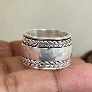 May include: A silver ring with a wide band and a braided design. The band is smooth and polished, and the braided design is raised and textured.