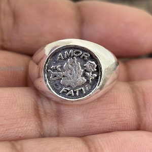 May include: A silver ring with a circular face. The face features the words "AMOR FATI" and a central design of a flame, a rose, and other symbols. The ring is round and has a polished finish.