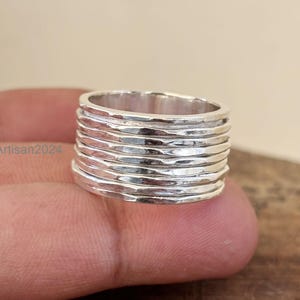 Könnte beinhalten: Ein silberner Ring mit einem strukturierten, geschichteten Design. Der Ring hat eine Reihe horizontaler Bänder, die ein einzigartiges Aussehen erzeugen. Der Ring wird von den Fingern einer Person gehalten.