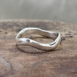 May include: A sterling silver ring with a wavy design. The ring has a smooth, polished finish and a flowing, organic shape. The ring is displayed on a wooden surface.
