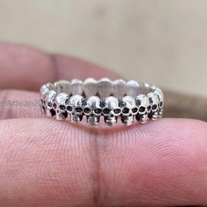 May include: A silver ring featuring a repeating pattern of small skulls. The ring is held up by a finger, showcasing the intricate details of the skull design. The ring is a gothic or alternative style.