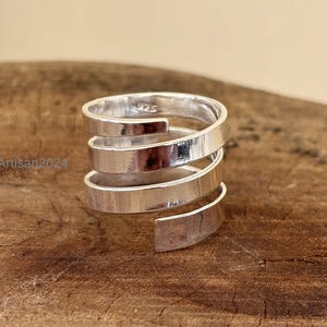 May include: A silver ring with a spiral design. The ring is made of polished metal and features a wrap-around style with multiple bands. The ring is set against a wooden surface.