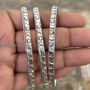Peut inclure: Trois bracelets argentés ornés d'un motif répétitif d'étoiles et de croissants de lune. Les bracelets sont tenus dans une main, mettant en valeur le design céleste. Les bijoux sont probablement en argent sterling.