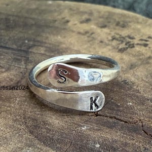 May include: A silver-colored bypass ring featuring a polished finish and two open ends. Each end is stamped with a single black letter, specifically the letter S on the top end and the letter K on the bottom end. The ring has a smooth, metallic surface.