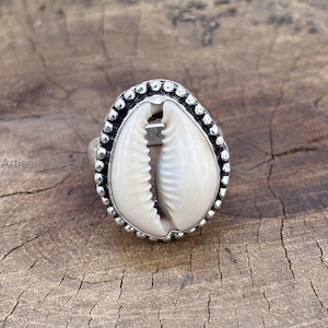 Puede incluir: Anillo de plata con una concha marina blanca. La concha ovalada está engastada en un borde plateado con cuentas. El anillo se muestra sobre una superficie de madera texturizada.