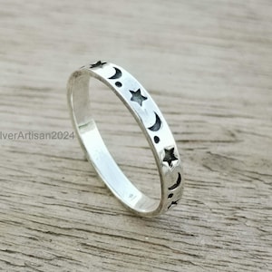 May include: A silver ring with a celestial design featuring stars, crescent moons, and dots. The ring is set against a wooden surface. The ring is a band style, with the celestial shapes cut out around the band.