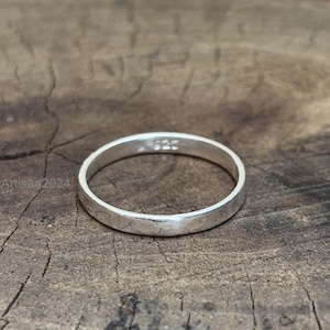 May include: A simple, silver ring with a smooth, polished surface. The ring is circular and appears to be made of metal. The number "925" is stamped on the ring. The ring is set against a rustic, wooden background.