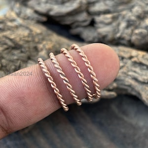 May include: Four copper-colored, twisted-rope style rings are displayed on a finger. The rings have a delicate, braided appearance. The background is a blurred view of a wooden surface.