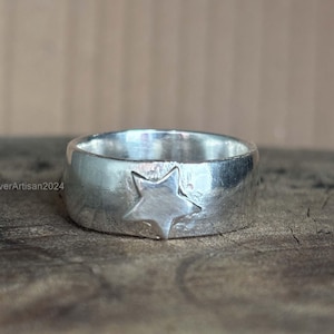 Può includere: Un anello d'argento con un design a stella. L'anello ha una fascia larga con una superficie strutturata e una stella in rilievo sul davanti. L'anello è esposto su una superficie di legno.