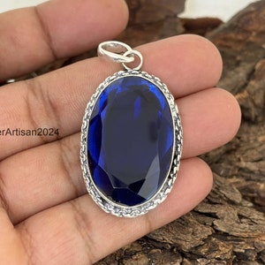 Può includere: Ciondolo ovale con una grande pietra preziosa blu scuro. La pietra è incastonata in una cornice di metallo color argento con un bordo decorativo a catena. Il ciondolo ha un piccolo anello nella parte superiore. Il testo "SilverArtisan2024" è visibile.