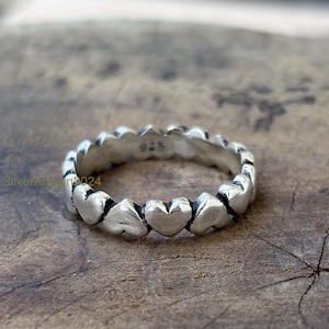 Peut inclure: Une bague en argent ornée d'un motif de cœurs répétés. La bague est en métal brillant et a un aspect texturé. La bague est posée sur une surface en bois. La bague est marquée "925".