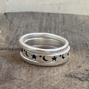 Puede incluir: Un anillo de plata con una banda que presenta un patrón repetitivo de estrellas y lunas crecientes negras grabadas. El anillo tiene un acabado liso y pulido, e incluye el texto SilverArtisan2024 visible en el lateral de la banda metálica.