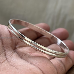 May include: A silver-colored bangle bracelet with a smooth, polished finish. The bracelet has a simple, elegant design, with a slightly rounded shape. The metal reflects light, creating a bright, shiny appearance. The bangle is a classic piece of jewelry.