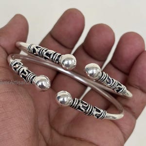 Op de afbeelding: Een set zilverkleurige, open armbanden. Elke armband heeft decoratieve zwart geëtste banden en gladde, ronde zilveren accenten. De sieraden worden in een hand getoond.