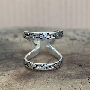 May include: A silver ring with a double-band design. The bands are connected by a curved support. Floral and vine patterns decorate the bands. The ring is displayed on a wooden surface. The text "SilverArtisan2024" is visible.