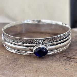 Pode incluir: Uma pulseira prateada com uma pedra preciosa oval azul escuro. A pulseira apresenta intrincados padrões florais e um acabamento polido. A pedra preciosa é engastada numa moldura, adicionando um toque de elegância à joia. Cerca de 2 cm de largura.