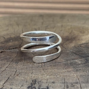 Op de afbeelding: Een zilveren spiraalring met een gehamerde textuur. De ring wikkelt zich in een doorlopend ontwerp om de vinger. De ring wordt gepresenteerd op een houten oppervlak.