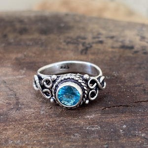 Puede incluir: Un anillo de plata de ley con una piedra preciosa redonda, facetada y de color azul brillante, engastada en un bisel con textura de cuerda. La banda muestra intrincados adornos y detalles oxidados, con el sello 925 en el interior.