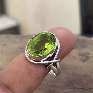 Może przedstawiać: Srebrny pierścionek z dużym, owalnym, jasnozielonym kamieniem perydot. Perydot jest osadzony w oprawie z lunetą, a pierścionek ma szczegółowy, ażurowy wzór na obrączce. Pierścionek jest prezentowany na palcu.