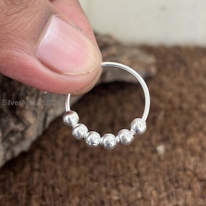 May include: A silver nose ring featuring six small, spherical beads. The ring is held by a person's fingers. The text "SilverArtisan2024" is visible on the ring. A simple and elegant piece of jewellery.