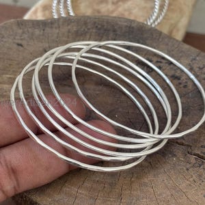 May include: A set of silver bangles with a hammered texture. The bangles are round and thin, with a bright, reflective surface. The image shows a stack of bangles, with a few more in the background. The bangles are displayed on a wooden surface.