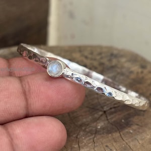 Könnte beinhalten: Ein silbernes Armreif-Armband mit gehämmertem Muster und einem kleinen, runden, hellblauen Mondstein. Das Armband ist schlicht und elegant, mit einem klassischen Design.