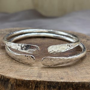 May include: A silver-colored, hammered-texture cuff bracelet. The open-ended design features a textured surface, reflecting light. The bracelet is displayed on a wooden surface, showcasing its handcrafted appearance. This piece is a stylish accessory.