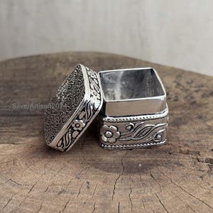 May include: A small, open silver box with a hinged lid, resting on a wooden surface. The lid is decorated with an intricate floral and scroll pattern. The box's body features a floral design. The box is square-shaped.