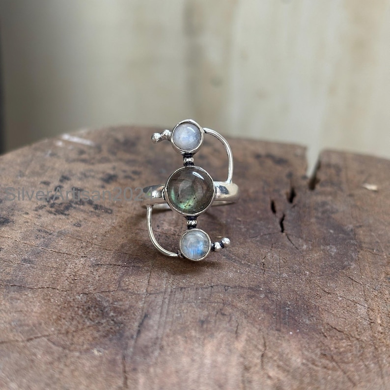 May include: A silver ring featuring three round gemstones. The central stone is a dark, translucent color, while the top and bottom stones are a lighter, opalescent hue. The ring has a unique, open design with curved silver accents.