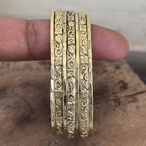 May include: Six gold-coloured bangles are stacked together. Each bangle has an intricate floral and leaf pattern. The bangles are displayed together, highlighting the detailed design. The surface of the bangles has a textured appearance, adding depth to the design.