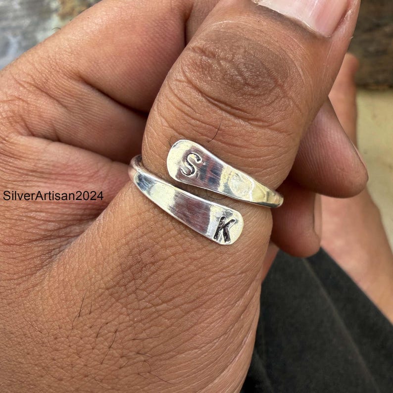 May include: A silver-colored bypass ring featuring two flat, tapered ends. One end is stamped with the letter S and the other end is stamped with the letter K. The metal has a polished, reflective finish and the band is open, allowing for adjustment.