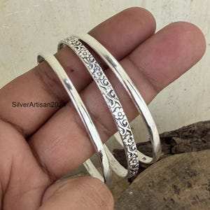 Peut inclure: Trois bracelets joncs en argent, comprenant deux anneaux lisses et polis et un anneau central orné d'un motif floral et végétal en relief. Ces bracelets métalliques, réfléchissants et circulaires, présentent une finition brillante.