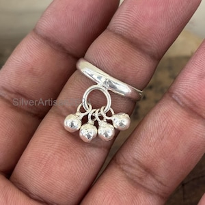 Könnte beinhalten: Ein Silberring mit einem Anhänger aus fünf kleinen, runden Glocken. Der Ring hat ein schlichtes Design mit glatter, polierter Oberfläche. Die Glocken sind an einem kleinen Ring befestigt, der am Hauptband hängt.