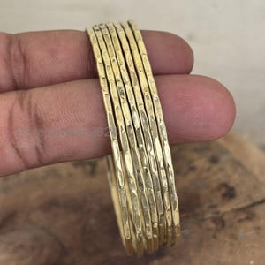 Op de afbeelding: Een stapel goudkleurige armbanden met een getextureerde, gehamerde afwerking. De armbanden zijn op elkaar gestapeld en tonen hun individuele ontwerpen en de algehele esthetiek. De sieraden worden in een hand gehouden.