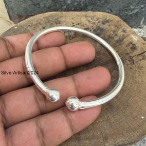 May include: A polished silver-tone open cuff bracelet featuring a smooth, rounded band that terminates in two spherical ball ends. The metal surface has a reflective, metallic finish, and the design is a simple, continuous circular shape.